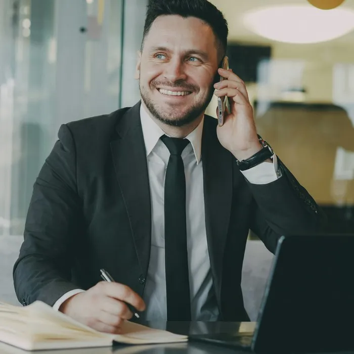 A smiling man in a suit on the phone at his desk